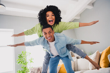 Photo of excited funky couple dressed casual outfits flying plane wings having fun indoors house...