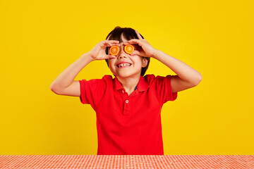Cheerful little boy in red polo shirt using carrot slices as pretend glasses against yellow...