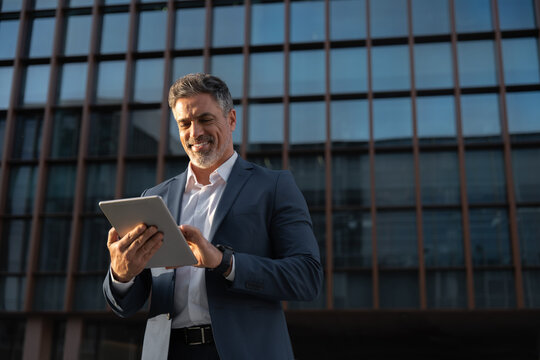 Focused successful mature Indian or Latin entrepreneur businessman holding digital pc tablet standing outdoor at business office building. Hispanic smiling man in suit working using touchpad computer