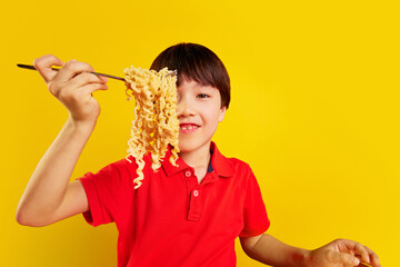 Smiling Korean boy, child in red polo shirt lifting noodles with fork against yellow background....