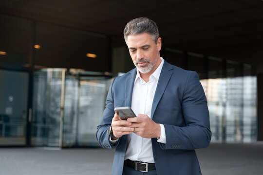 Successful mid age senior businessman holding mobile phone. Portrait of focused executive ceo manager. Latin adult man in suit using smartphone application app device for business at office outdoors