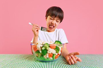 Little Korean boy sitting at table with fork and eating boiled vegetables from a bowl against pink background. Broccoli, carrots, and cauliflower. Concept of food, healthy eating, childhood, pop art