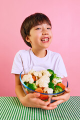 Delightful mealtime. Smiling boy, child sitting at table with bowl full of boiled broccoli, carrots, and cauliflower over pink background. Concept of food, healthy eating, childhood, emotions, pop art