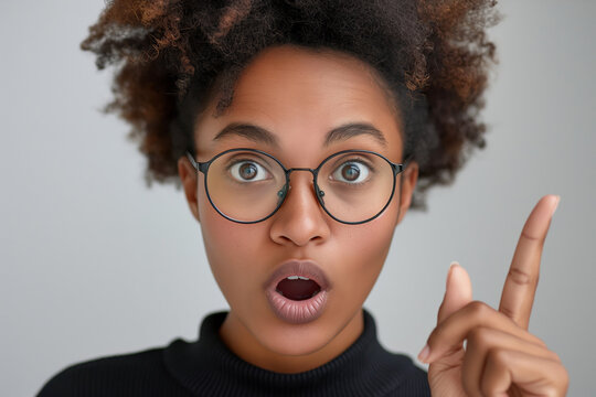 Surprised young woman wearing glasses, pointing upwards. Ideal for depicting moments of realization in ads, presentations and educational content