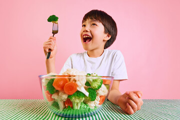 Playful and healthy. Smiling boy, kid raising for with broccoli, sitting at table with bowl full of boiled vegetables on pink background. Concept of food, healthy eating, childhood, emotions, pop art