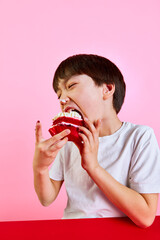 Moment of happiness. Boy, child enjoying delicious red velvet cake with cream and berries, making big bite against pink background. Concept of food, childhood, emotions, meal, menu, pop art