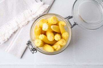 Tasty fresh yellow baby corns in glass jar on white tiled table, top view