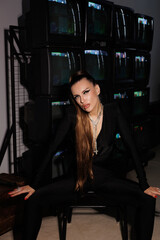
A beautiful girl in a black jumpsuit with an open neckline and high heels, against the backdrop of old working TVs standing in a row. A portrait of girl with bright makeup, winged eyes, big lips.