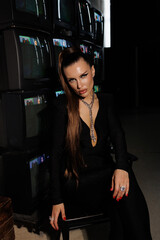
A beautiful girl in a black jumpsuit with an open neckline and high heels, against the backdrop of old working TVs standing in a row. A portrait of girl with bright makeup, winged eyes, big lips.