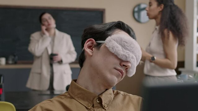 Tilt up of young man wearing sleeping mask sitting at desk and napping during workday in modern office while his colleagues resting playing table tennis in blurred background