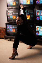 A beautiful girl in a black jumpsuit with an open neckline and high heels, against the backdrop of old working TVs standing in a row. A portrait of girl with bright makeup, winged eyes, big lips.