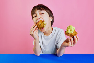 Moment of pleasure. Little boy, child in white t-shirt sitting at table and emotionally eating fried chicken with mustard sauce against pink background. Concept of food, childhood, emotions, pop art