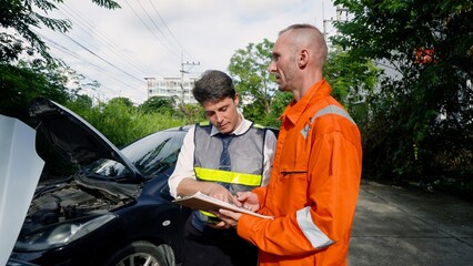 Two men, one in safety vest and another in orange work suit, examining black car engine, notepad in...