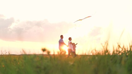 Cute little girl with flying kite mother father holding hands walking at sunset field together. Happy family parents and daughter outdoor playing flight toy entertainment having fun at nature meadow