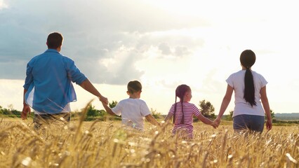 Family holding hands walking at sunset sunny dry wheat field outdoor weekend back view. Parents and children going together natural sunlight agriculture countryside dramatic sky scenery enjoy freedom