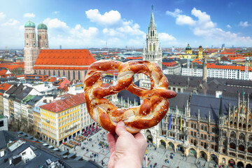 A hand is holding a traditional German Pretzel in front of the skyline of Munich, Germany, as a...
