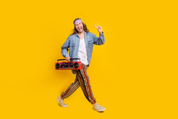Full size profile photo of cool young man boombox show v-sign walk empty space wear denim shirt isolated on yellow color background
