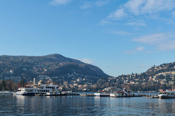 Beautiful view of floating boats and ships reflecting in the clean clear water of the lake of Como, against Italian Alps and blue sky background on a cold sunny winter day. Tourism and travel concept