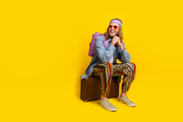 Full size photo of cool young man sit valise look empty space wear denim shirt isolated on yellow color background