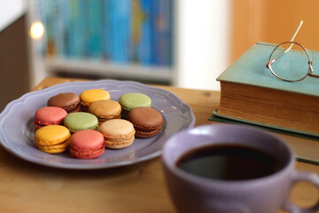 Purple plate filled with pastel macarons, cup of tea or coffee, vintage books and reading glasses on the table. Colorful bookcase in the background. Selective focus.