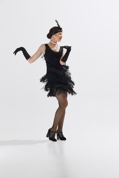 Beautiful young woman in vintage 1920s flapper dress, complete with fringes and feathered headband, dancing isolated over white studio background. Concept of art, retro and vintage, entertainment, 20s