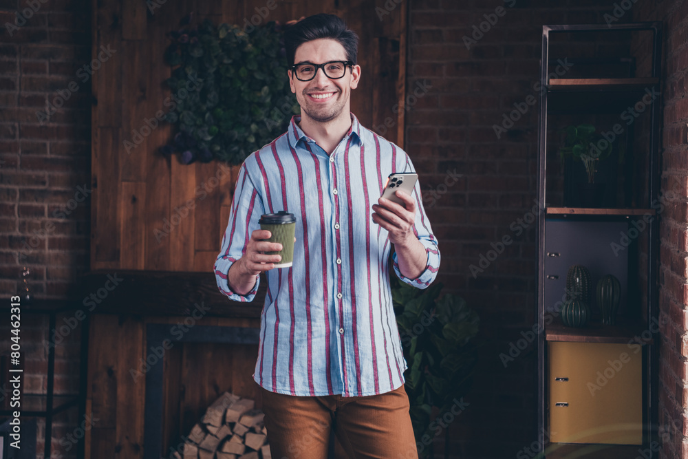 Poster Portrait of nice young man drink coffee use phone wear striped shirt loft interior business center indoors