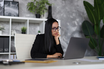 A pensive Asian businesswoman at her desk, contemplating a problem while working on her laptop.