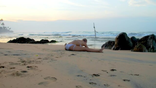 Woman athlete in bikini stretches body practicing leg-split on beach by ocean. Lady enjoys yoga session with view of crashing waves