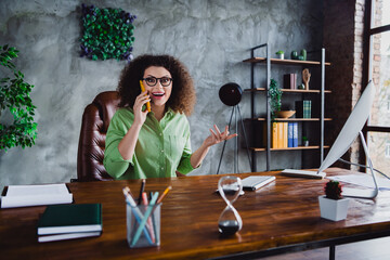 Photo of charming glad cheerful woman with curly brown hair sitting speaking phone partners dialog...