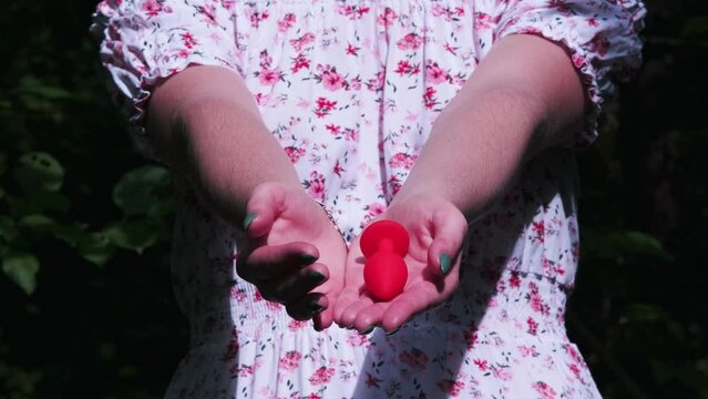 Close-up of a girl's hands in a delicate white summer dress. She opens palms, which contain a red butt plug. Sex toys, Sex shop. BDSM - outfit for adult sex games
