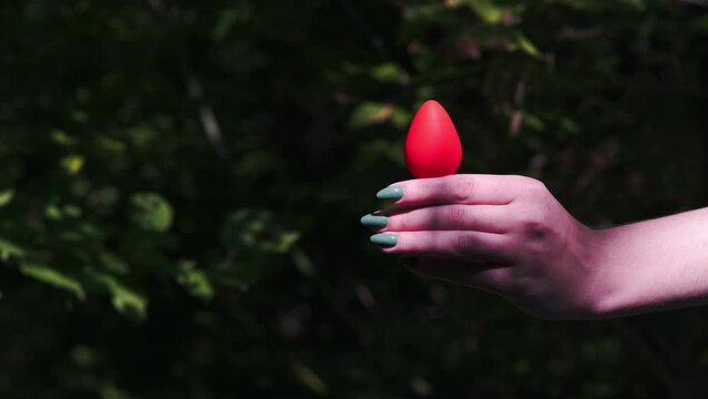 Woman's hand holds a red anal plug against a background of green bushes in the forest. Sex toys, Sex shop. BDSM - outfit for sex games for adults
