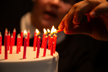 Detalhe de uma mão acendendo várias velas sobre um bolo de aniversário com um rosto desfocado ao fundo.