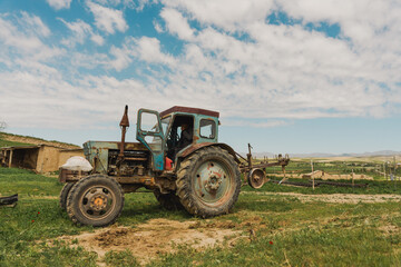 An old blue tractor is parked in a field. The tractor is rusty and has a worn-out appearance. The sky is clear, and the sun is shining brightly. The scene gives off a sense of nostalgia