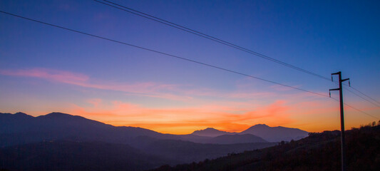 Blurred sunset behind mountains with power lines against cloudy sky