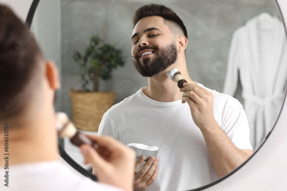 Wall mural Handsome young man shaving beard near mirror in bathroom