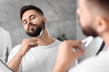 Handsome young man trimming beard with scissors near mirror in bathroom
