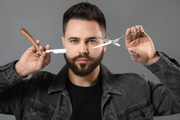 Handsome young man with mustache holding blade and scissors on grey background