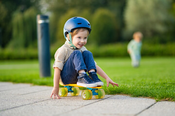 Cute little boy learning to skateboard on beautiful summer day in a park. Child wearing safety helmet enjoying skateboarding ride outdoors.
