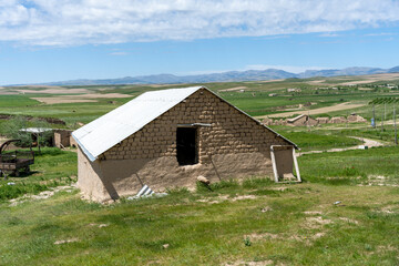 A small house with a white roof sits in a grassy field. The house is surrounded by a dirt road and a few other buildings. The sky is clear and the sun is shining brightly