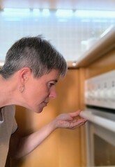 Profile of young adult woman, in front of the oven at home looking inside. Housewife checking the cooking. Housekeeper cooking.  Observe and check the cooking of the food