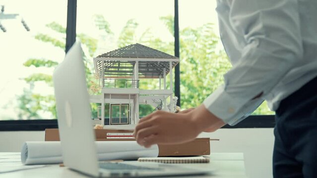 Close up of professional male architect engineer measure house model and using laptop analysis data at meeting table with house model and map . scatter around. Focus on hand. Immaculate.