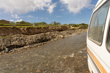 A white van is driving down a river with a cliff in the background. The water is choppy and the van...