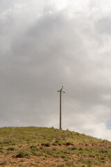 A tall pole with wires on it is standing on a hill. The sky is cloudy and the weather is overcast