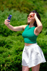 Beautiful girl in a green top and white shorts walks in a city park on a sunny summer day. Taking a selfie, laughing while holding a mobile phone - obrazy, fototapety, plakaty