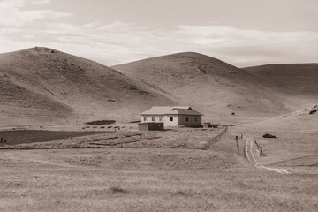 A small house sits in the middle of a vast, empty field. The house is surrounded by a dirt road and a fence. The sky is cloudy, and the sun is low in the sky. The scene is quiet and peaceful