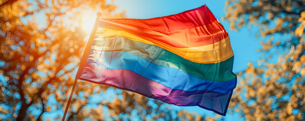 A rainbow flag is flying in the sky. The flag is colorful and vibrant, and it is a symbol of pride and acceptance. The flag is held up by a pole, and it is against a backdrop of trees and a blue sky