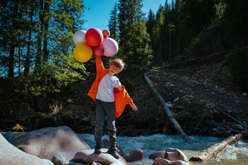Happy boy with balloons stands on the bank of mountain river