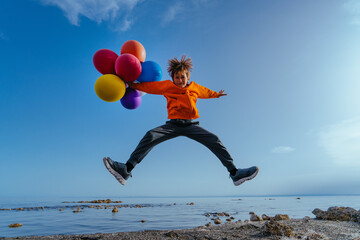 Cheerful boy jumping with balloons on lake shore on summer day