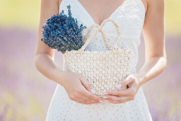 woman in white dress standing and holding straw bag with lavender flowers in her hand. Woman in...