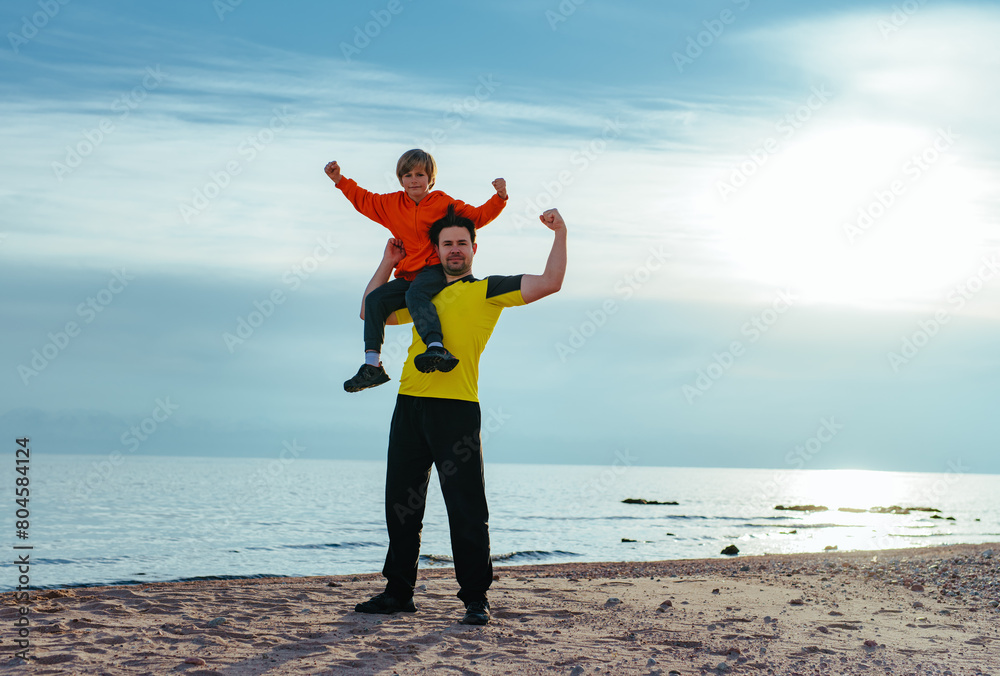 Poster Happy boy and his father showing muscles on lake shore at summer, boy sitting on father's shoulder